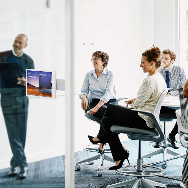 Modern conference room with a long rectangular table surrounded by chairs, a wall‐mounted display at one end showing scheduling panels, and large floor‑to‑ceiling windows letting in daylight